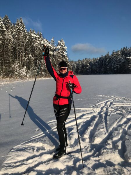A woman in a red jacket and black pants is standing in a snowy landscape on skis, holding up one of her ski sticks and smiling widely.