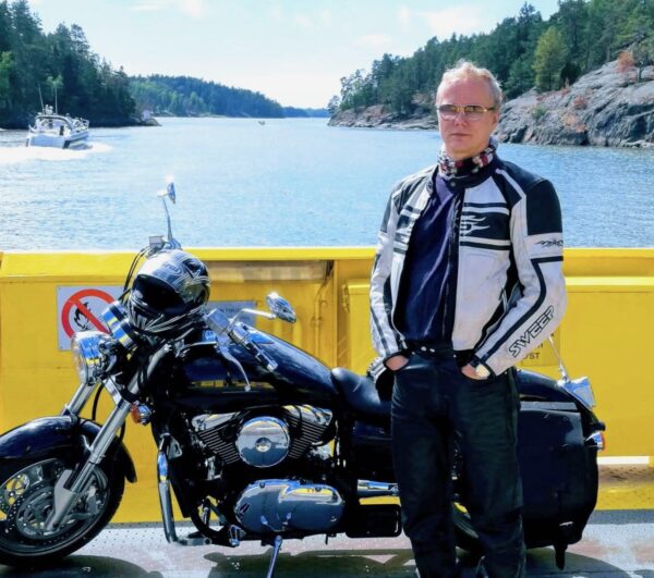 A man is standing in front of a motorcycle on a ferry, with the sea and islands in the background.