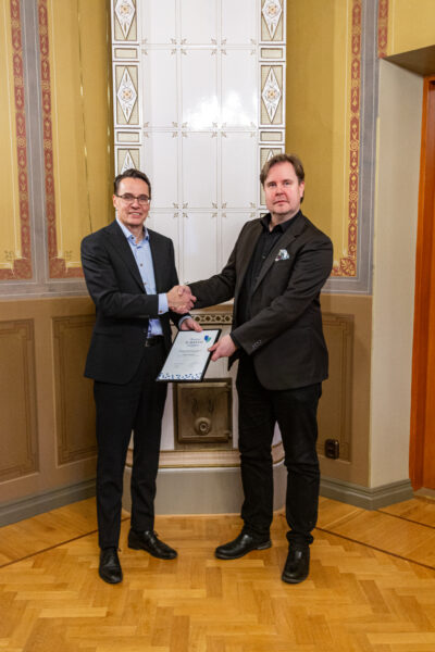 Two men in suits shaking hands and holding onto a plaque.