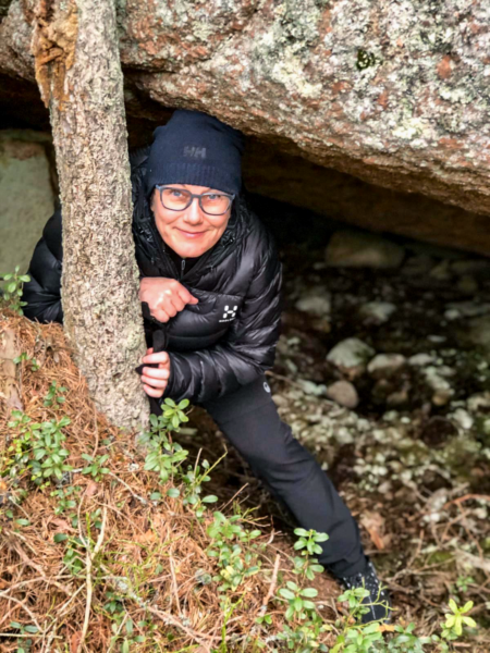 A woman stands underneath a large rock and smiles at the camera.