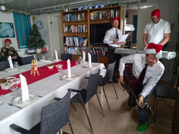 Men clad in suits and elf hats play music. There is a decorated table and a smiling woman sitting in the background.