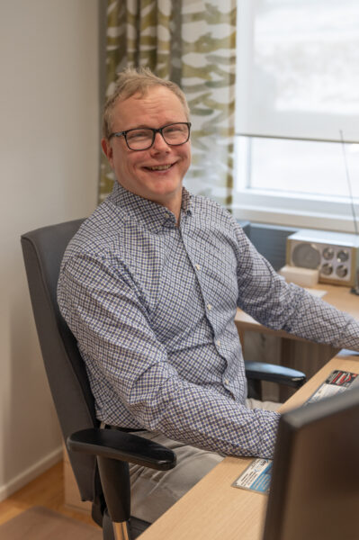 A blonde man in glasses is sitting behind a desk and smiling widely.