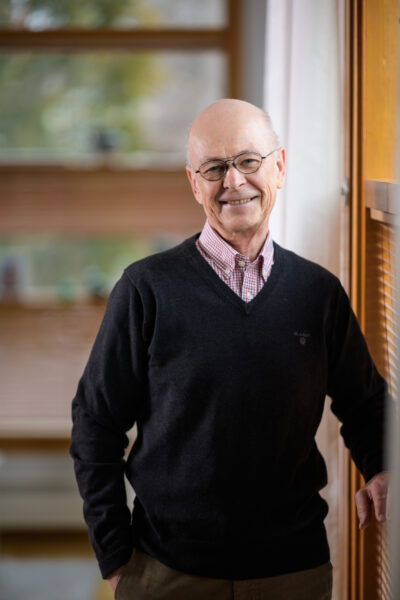 A man in glasses is smiling at the camera and leans against a window on the right.