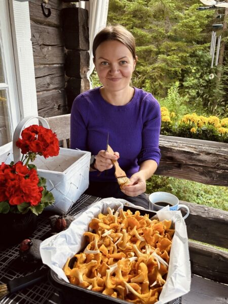 A woman in a purple shirt is sitting behind a table outdoors. On the table there is a red flower and a big container filled with chantarelles.