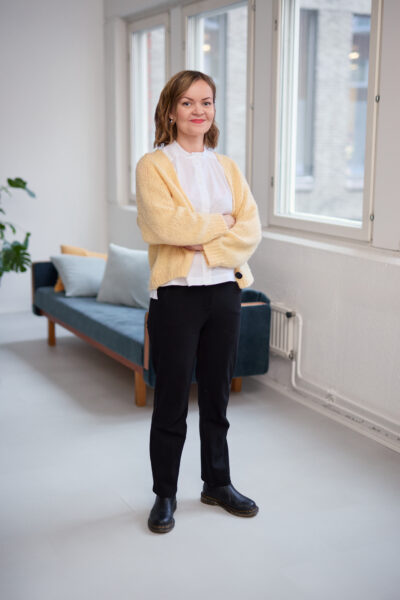 A woman in a yellow cardigan, white shirt and black pants is standing with her arms crossed in the middle of a room.