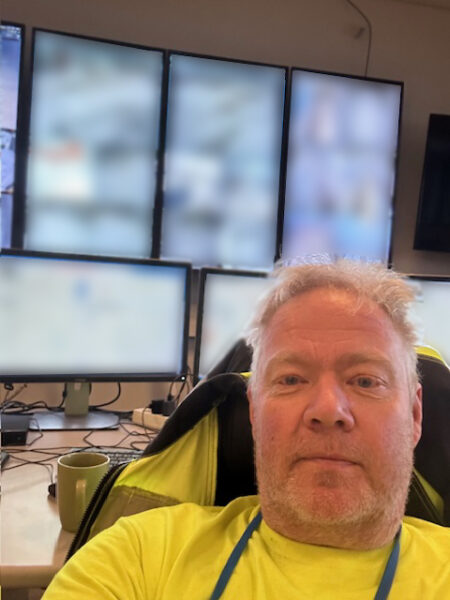 A man in a yellow shirt sitting in front of some blurred monitors.
