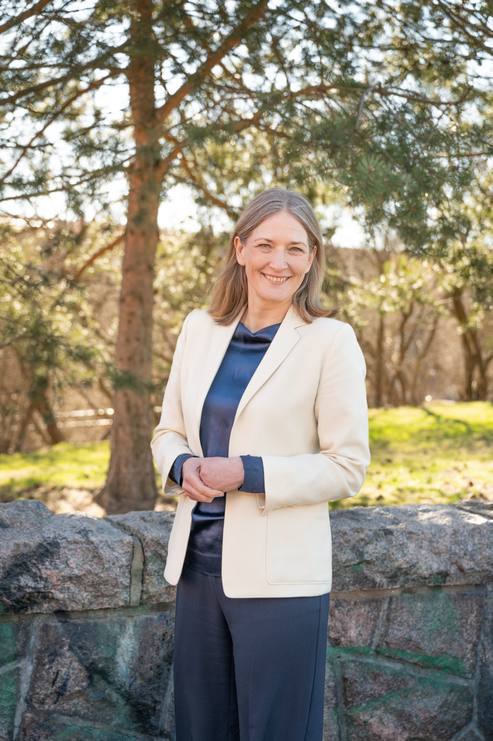A woman with a blue shirt and white blazer stands outside and smiles at the camera.