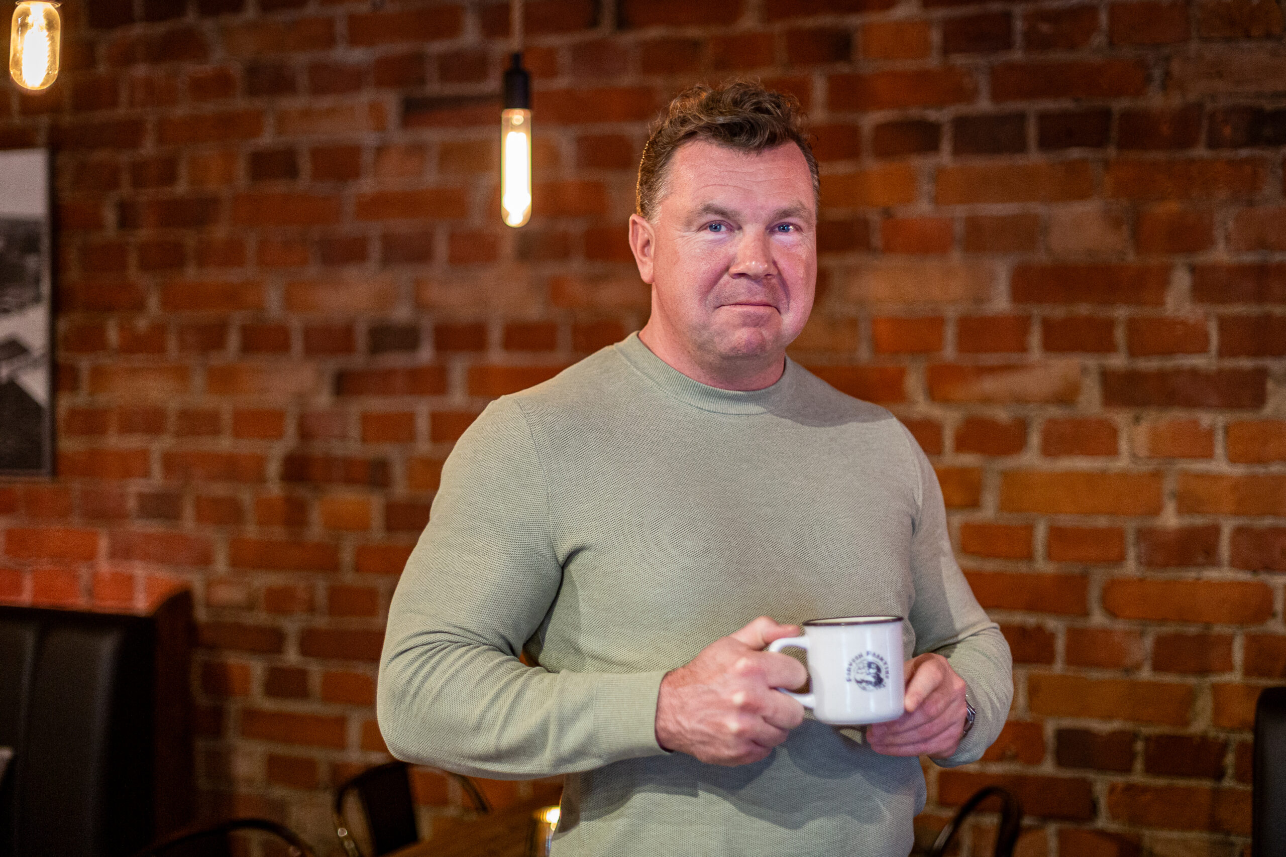 A man holding a coffee cup, standing in front of a red brick wall.
