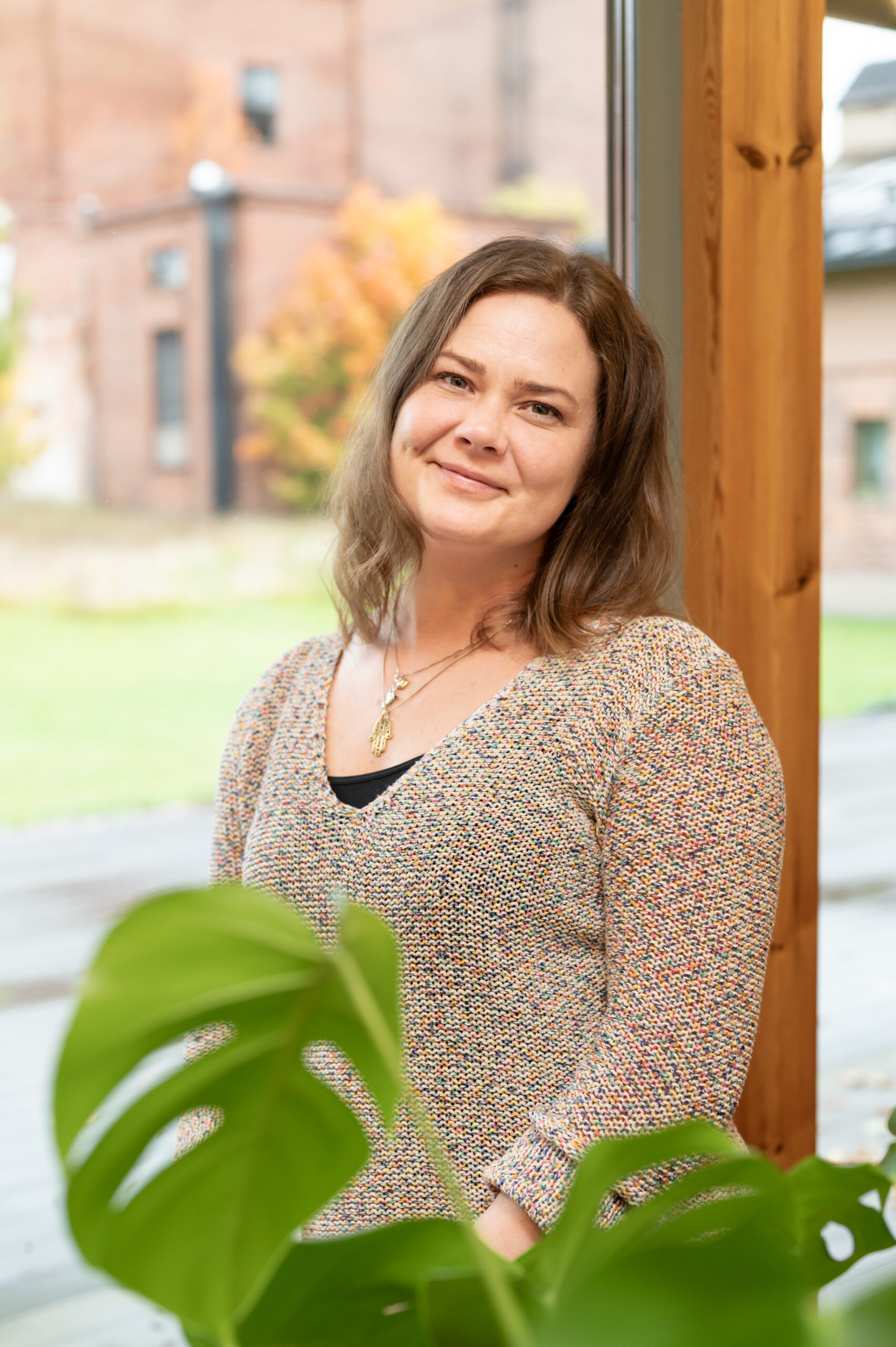 A woman is leaning back against a window and looking at the camera. A monstera plant in the foreground.