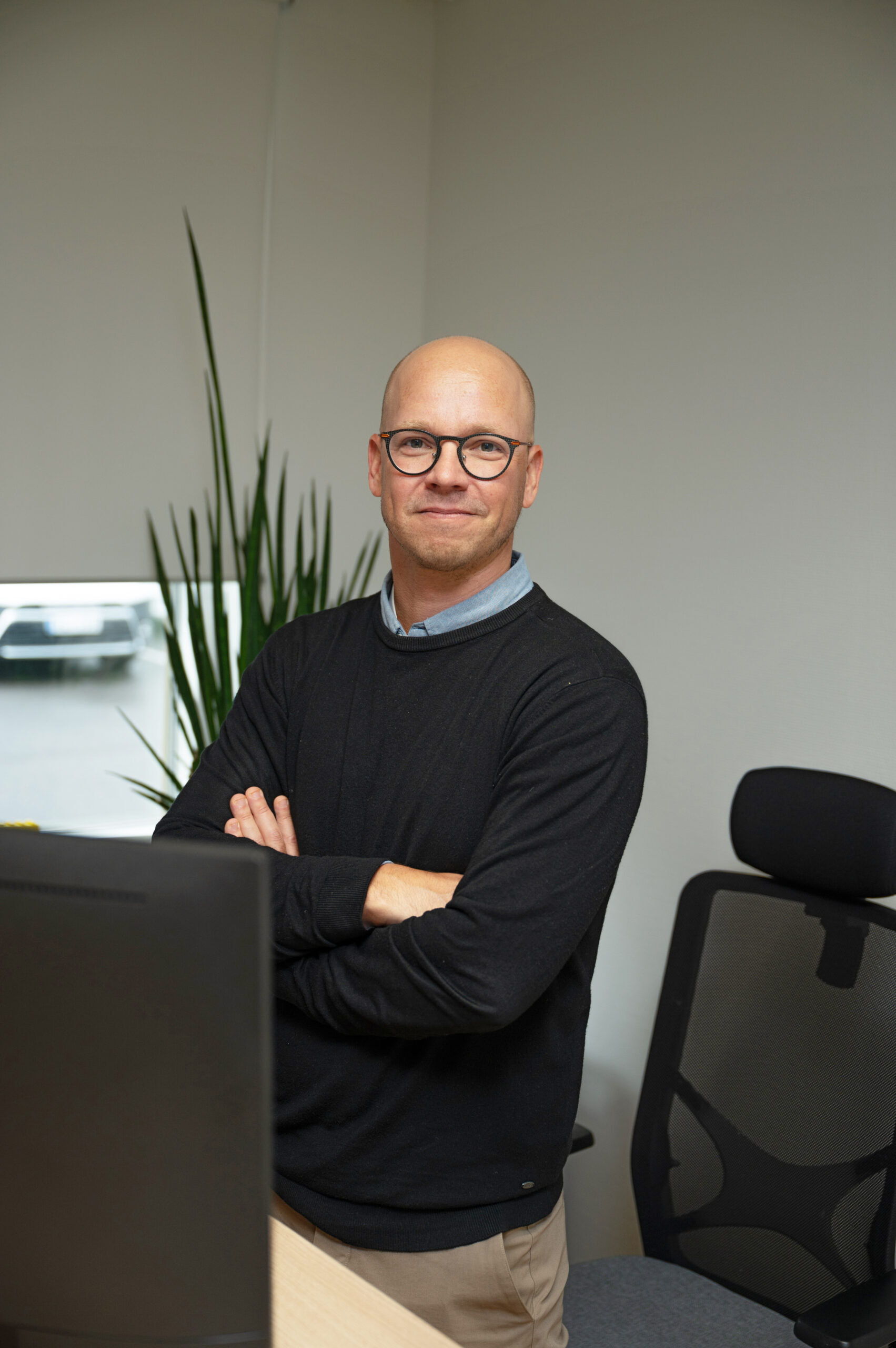A bald man with glasses is standing with his arms crossed behind a desk.