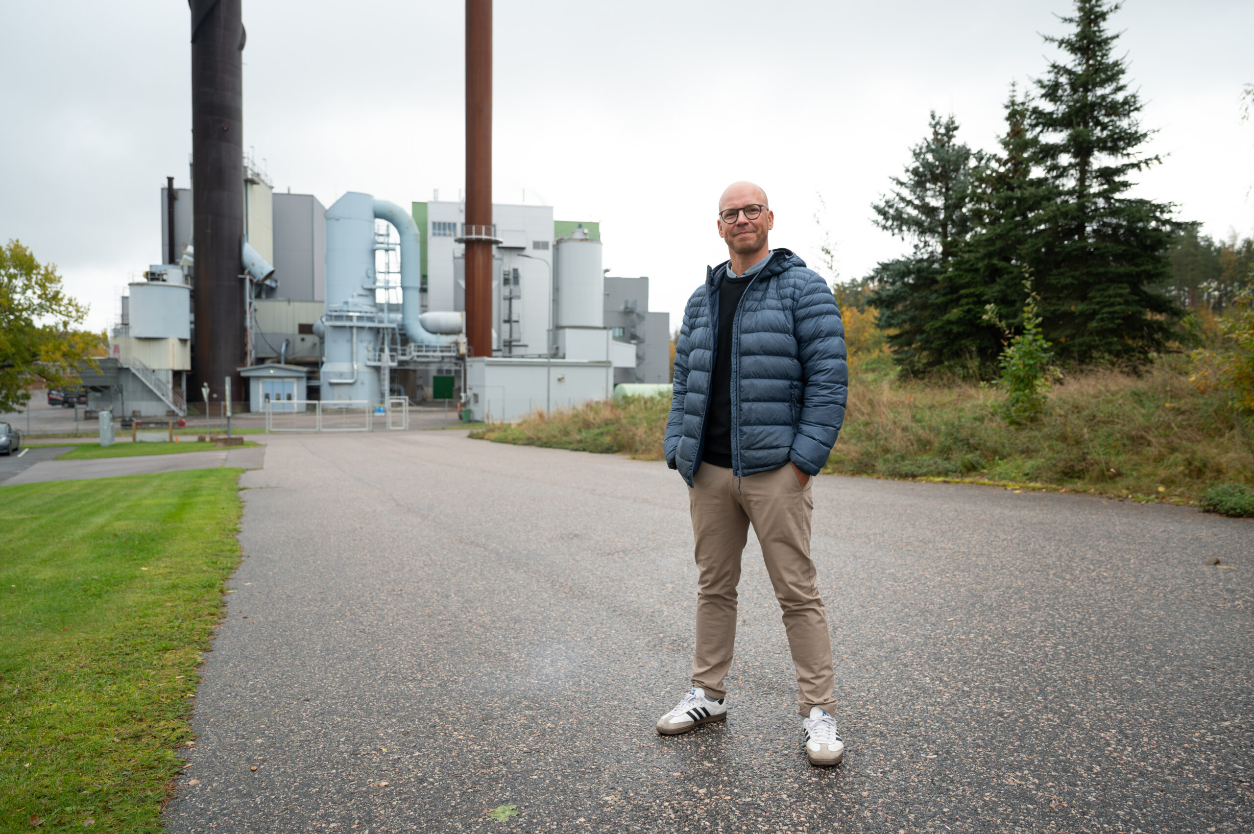 A bald man in glasses is standing with his hands in his pockets. In the background a powerplant.