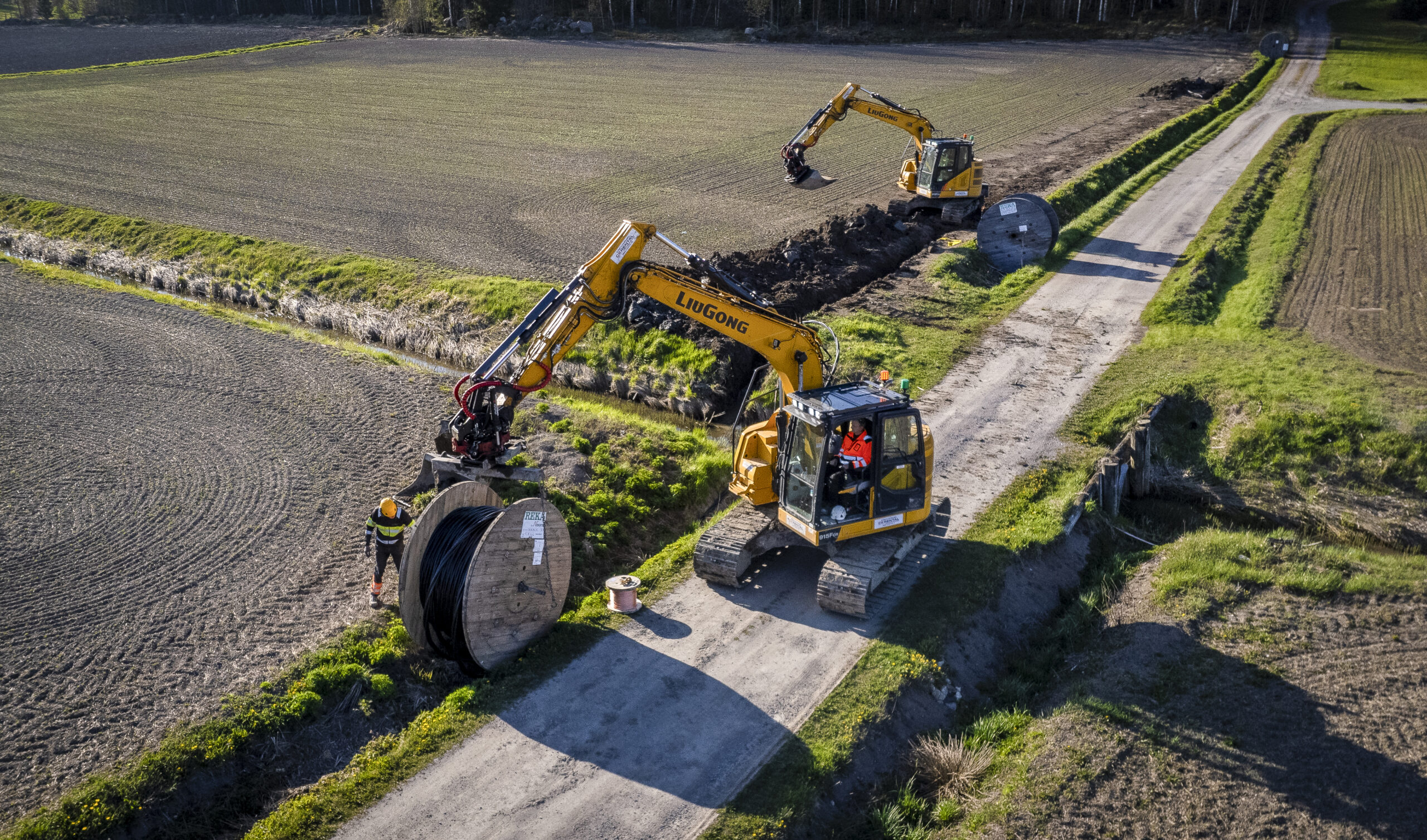 A country road and field photographed from above, diggers are putting electric cables in the ground.