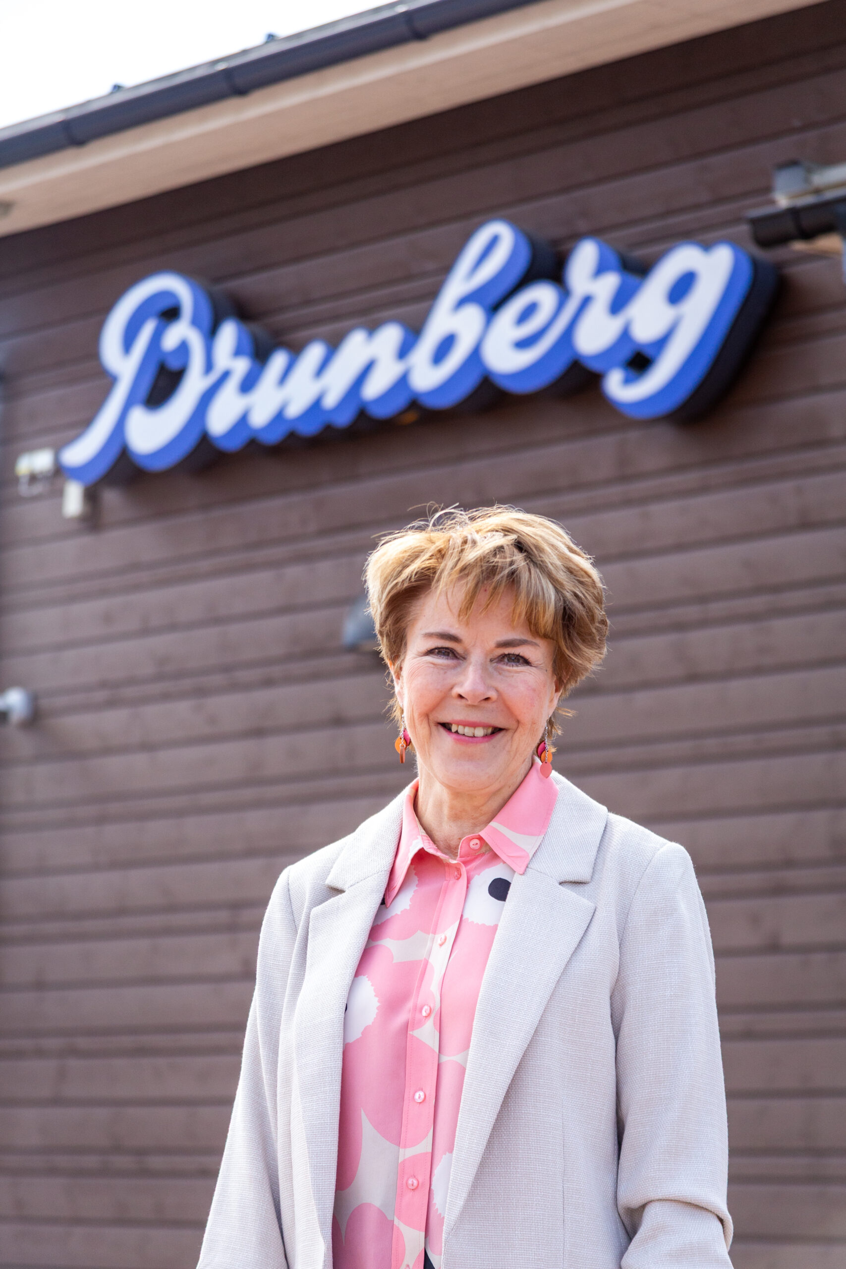 A woman in a white blazer and shirt with pink and white flowers stands in front of a wooden wall. On the wall is a sign that says "Brunberg."