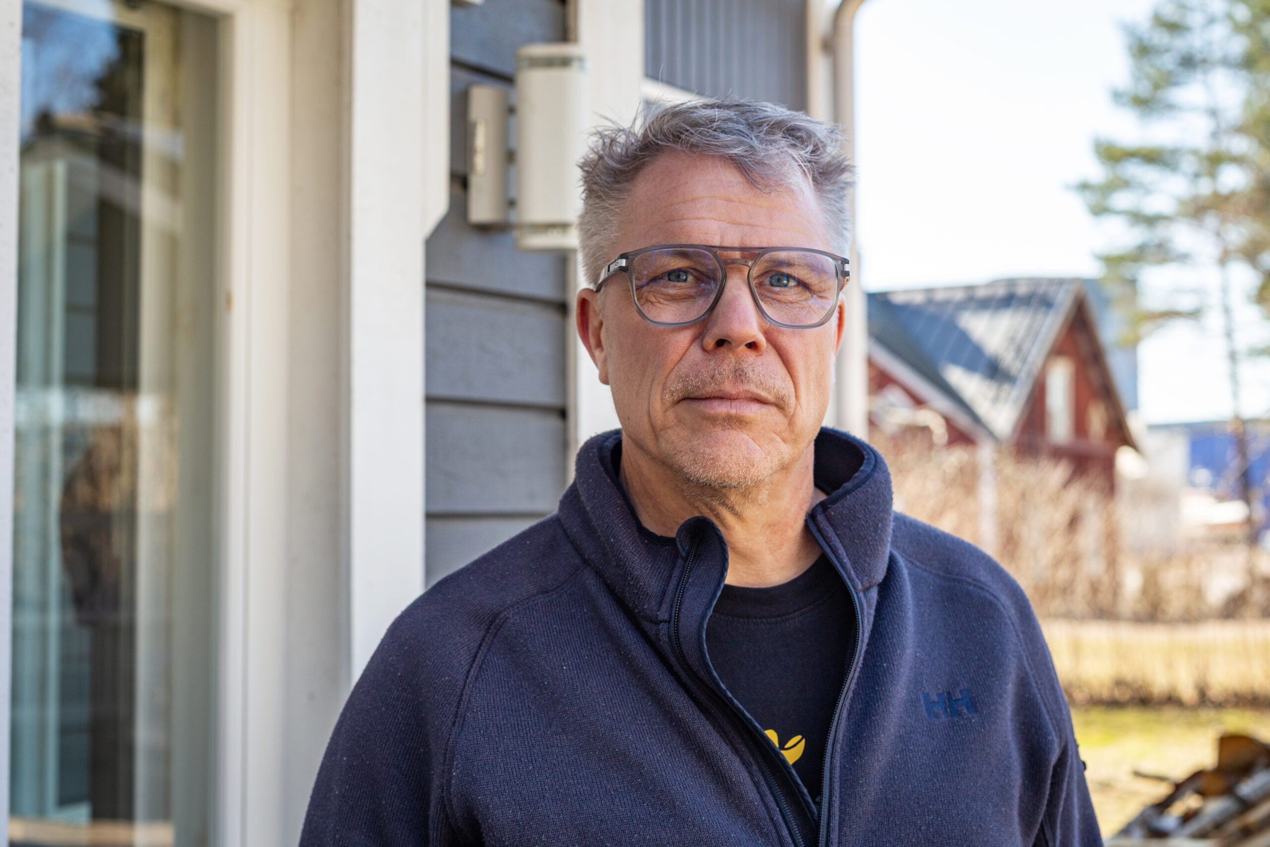 A man with grey hair, glasses and a blue shirt is posing in front of a house.