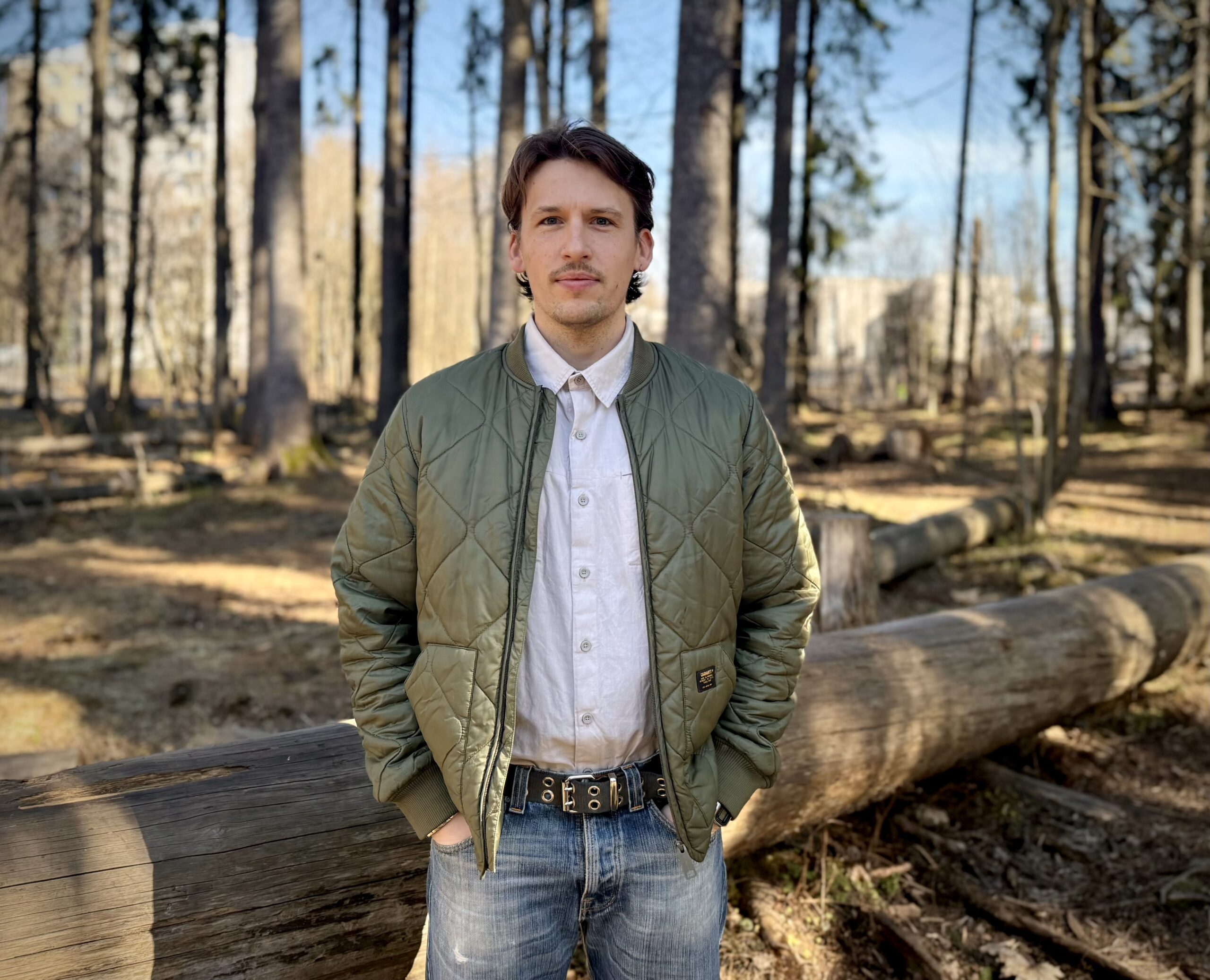 A man with his hands in his pockets is standing in a forest clearing.